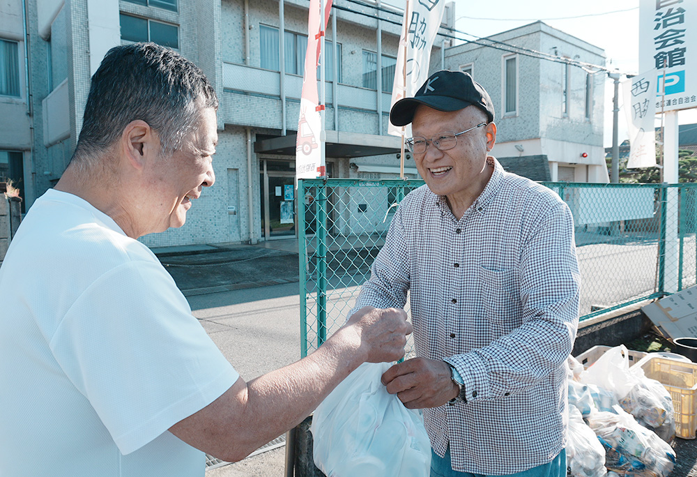 近所の人からアルミ缶を受け取る松本さん㊨